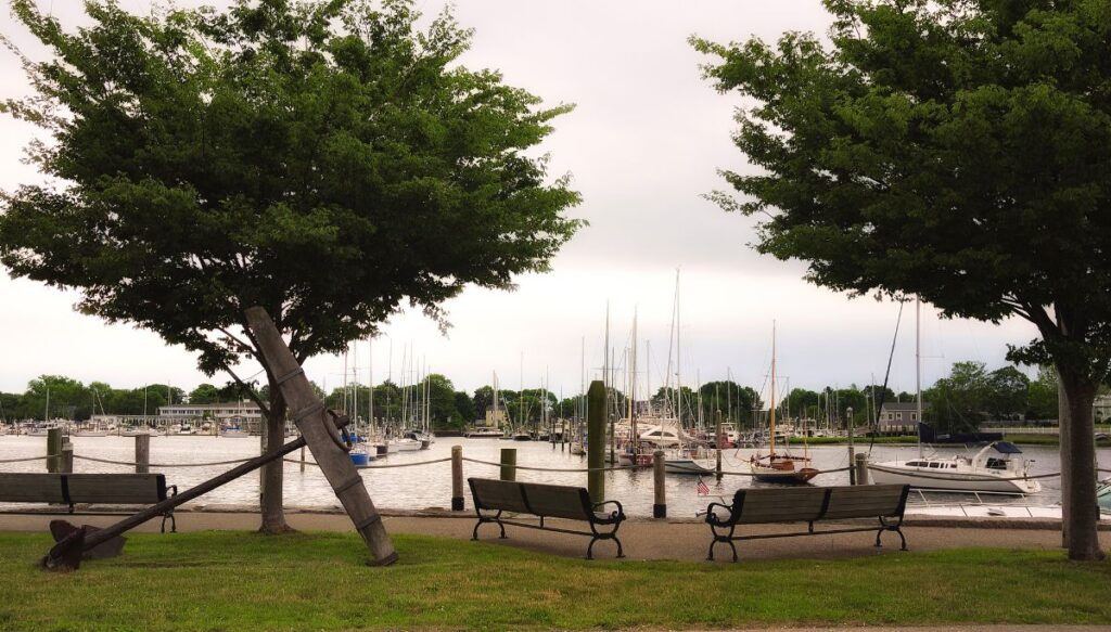 View of a path along the water with benches beside it and trees in the background. You can also see boats in the water to the front of the path.