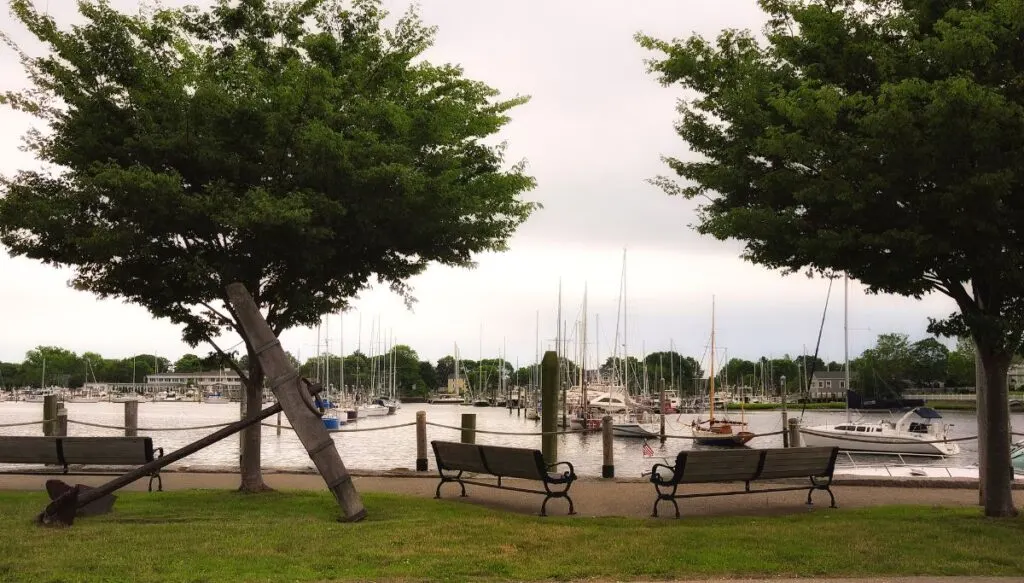 View of a path along the water with benches beside it and trees in the background. You can also see boats in the water to the front of the path. 