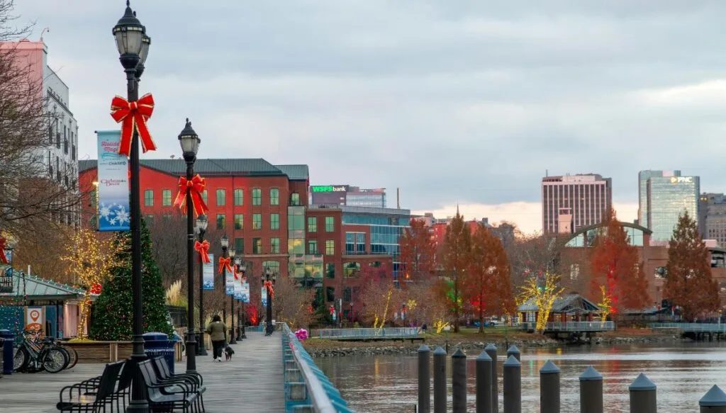 View of the Wilmington waterfront on a gray day with tons of holiday decor and people walking along the boardwalk on the river with tall buildings in the background. 