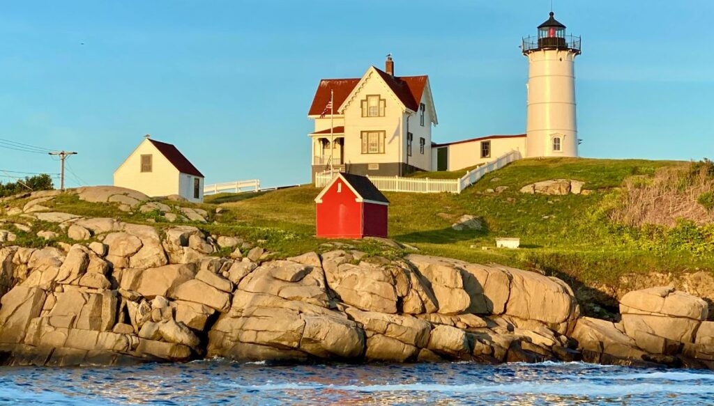 View of Nubble lighthouse sitting on top of a hill with rocks beneath it that lead to the ocean.