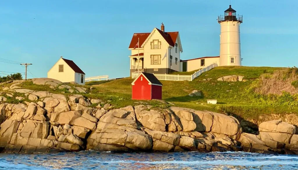 View of Nubble lighthouse sitting on top of a hill with rocks beneath it that lead to the ocean. 