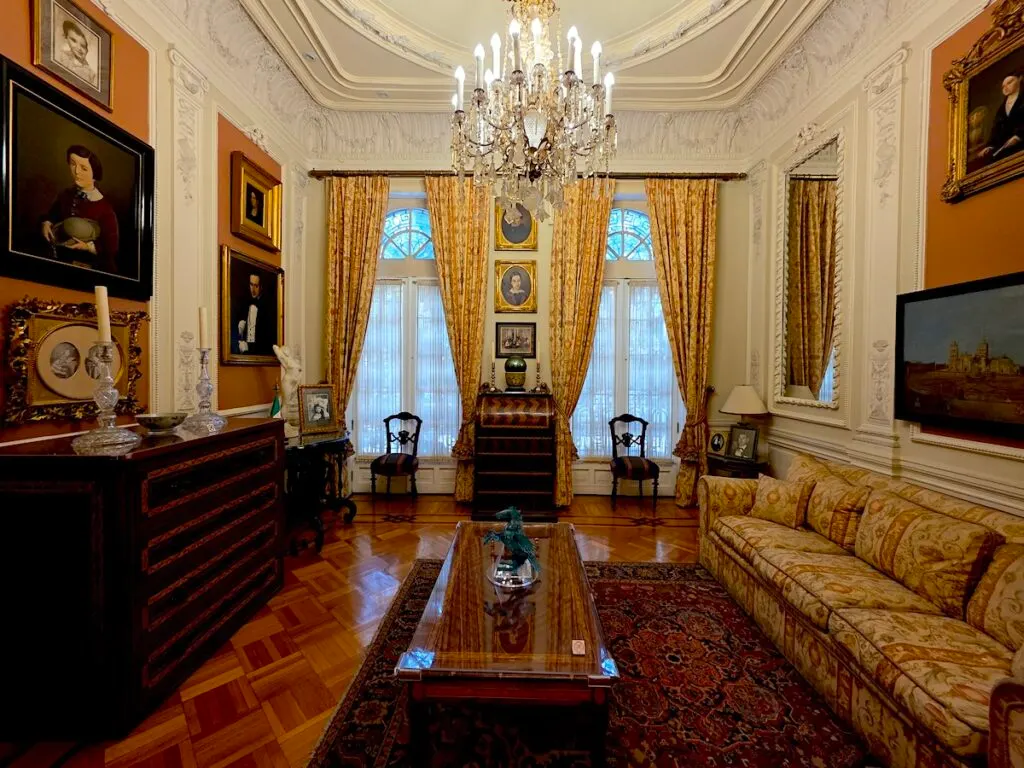 View of a colonial-style living room with yellow curtains, vintage portraits, a yellow couch, and a wooden table in the center. 