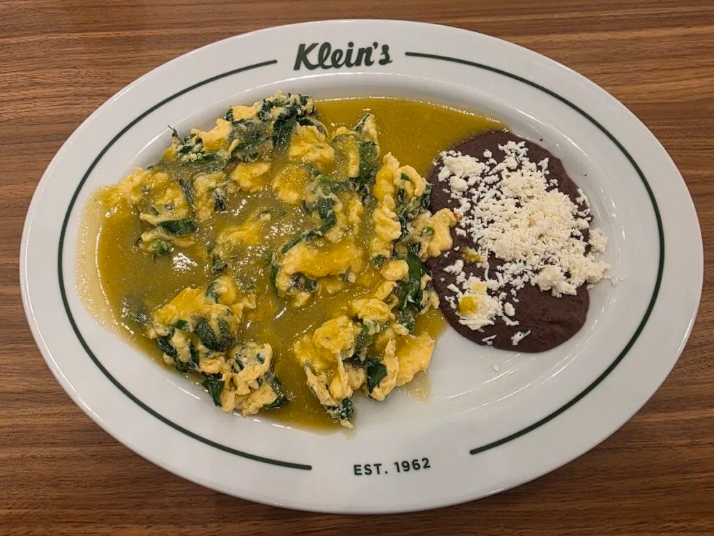 Aerial view of scrambled eggs with green sauce and spinach with black beans on the side and topped with cheese. 