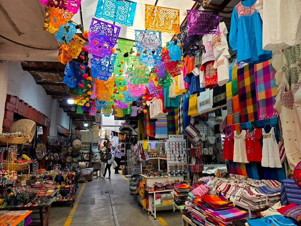 View of colorful flags hanging in the air and walkways filled with clothes and goods for sale inside Mercado de Artesanias La Ciudadela.