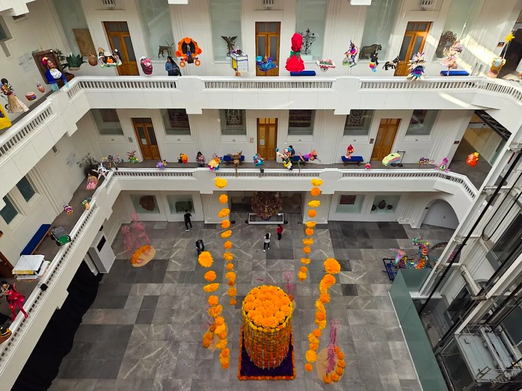 Aerial view of the atrium in Museo de Arte Popular. You can see three white floors with pi&ntilde;atas hanging and a large tower in the middle made of ,arigolds. 