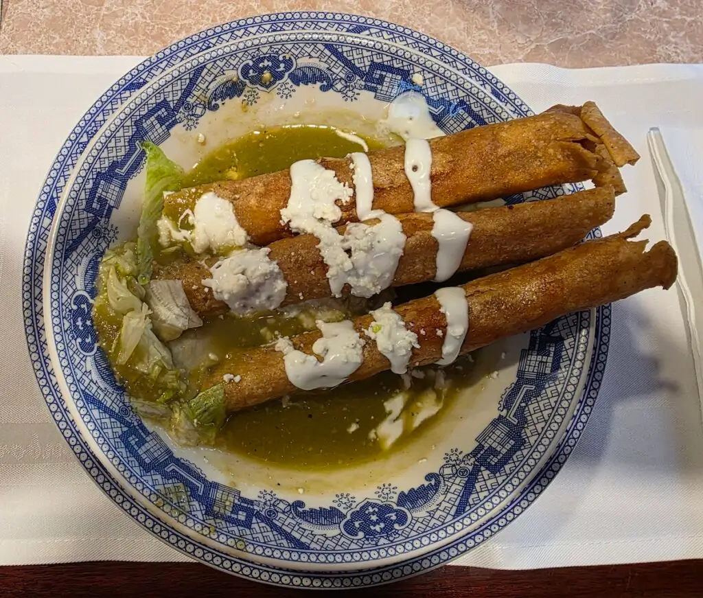 Aerial view of a plate with three fried flautas with a white a white sauce on top and a green sauce on the bottom of a blue ceramic plate. 