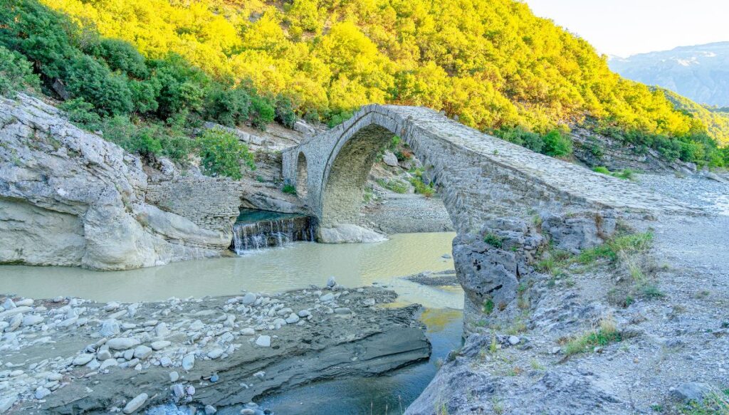 View of an arched stoned bridge going over water from the Benja Thermal Springs with trees glowing yellow in the sun behind the bridge.