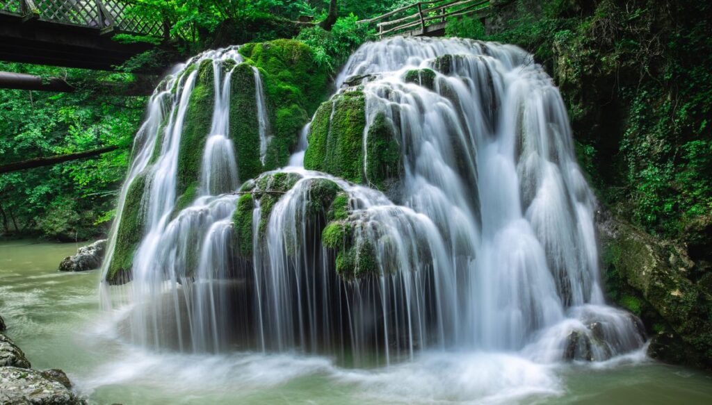 View of a waterfall streaming down moss covered rocks with a bridge above on the left at Bigăr Waterfall.
