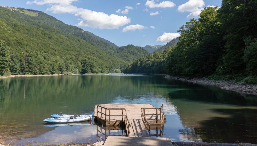 View of wooden pier with two white kayaks on the left in a lake that is surrounded by hills filled with green trees in  Biogradska Gora National Park. 