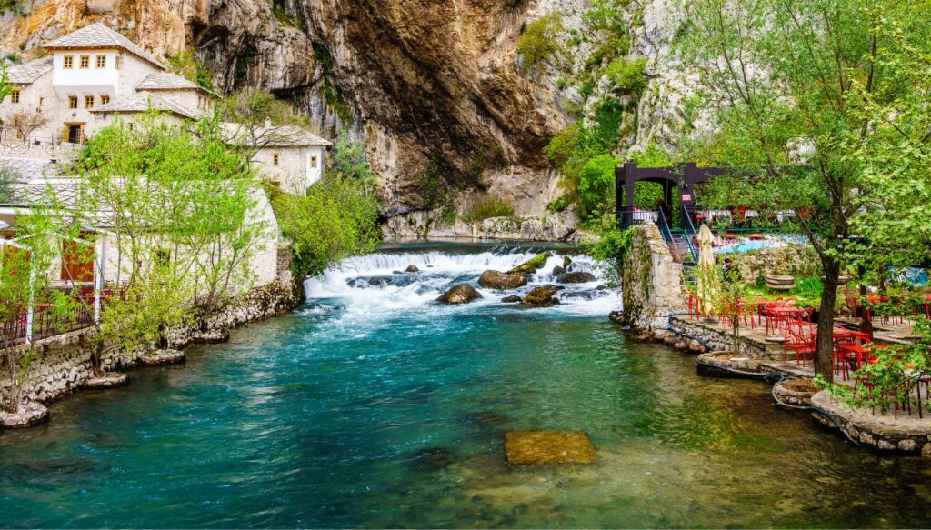 View of Blagaj Tekke on the left and a river with bright blue water in the middle with a stone mountain in the background.