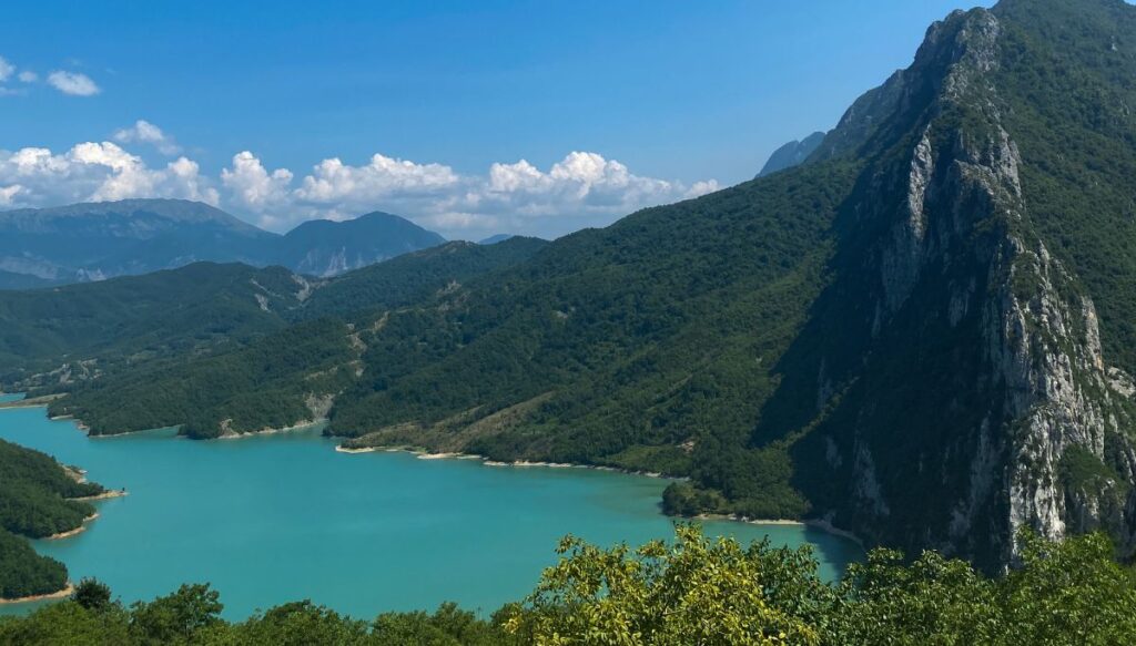 View of sheer mountains covered in forests surrounding a turquoise blue lake on a sunny day in Albania. This is Bovilla Lake.