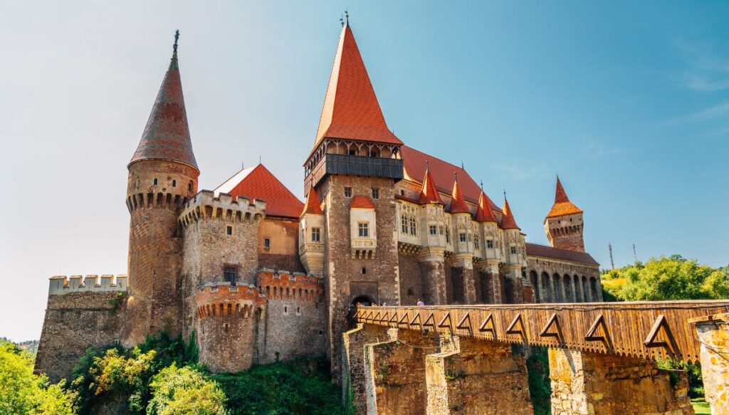 View of a medieval castle with a red roof and a wooden bridge leading up to the stone caste. This is Corvin Castle and it is easily one of the most beautiful places in Romania.