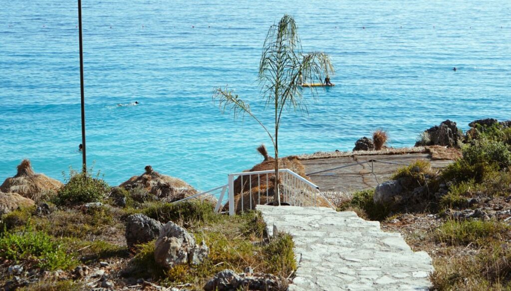 View of a stone path taking you to the edge of the water in Dhërmi in Albania with a talk tree and a man in a yellow boat in the water.