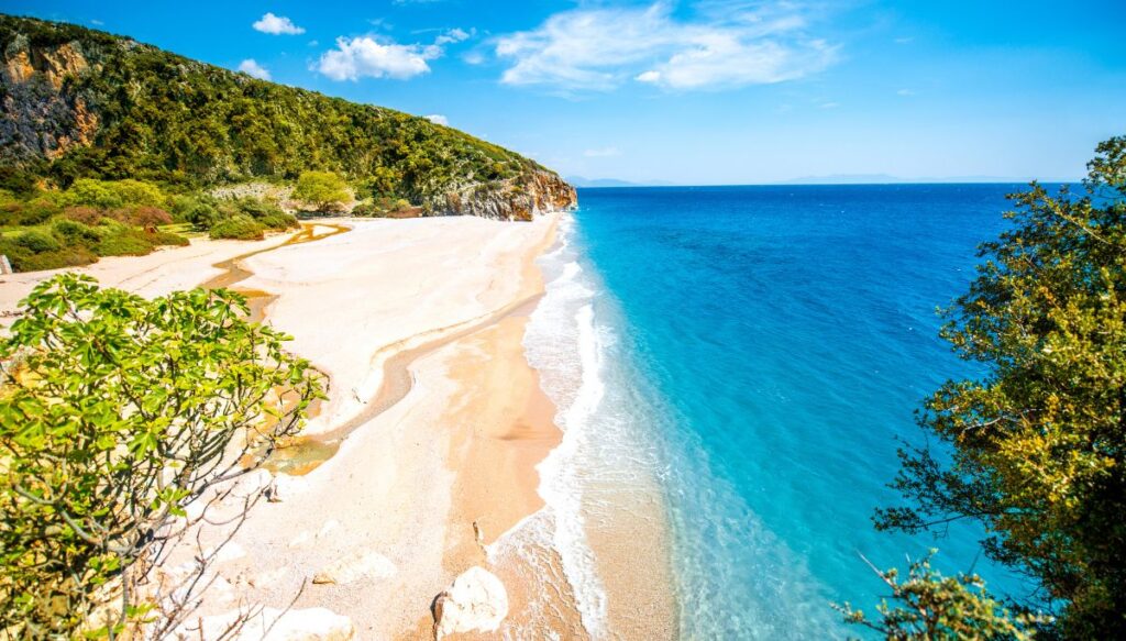 Aerial view of the white sand, the blue water, and the the forested hills surrounding Gjipe Beach.