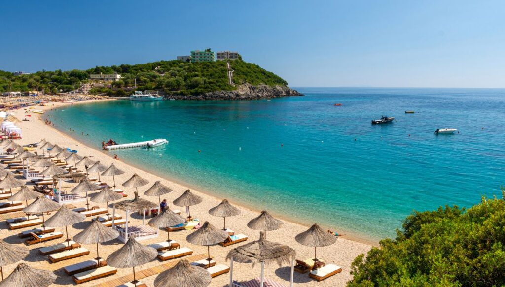 View of white long chairs under thatched umbrellas along the beach with boats in the water and a small hill covered in greenery and buildings in the distance on the coast in Himare.