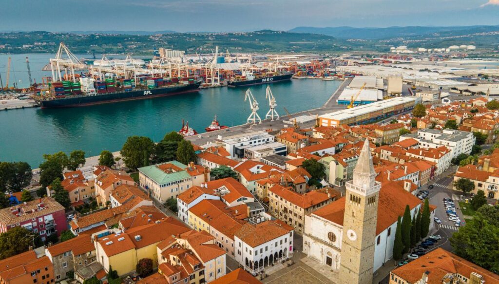 View of the church steeple and red roofed buildings along the sea in Koper with the port in the background of this aerial shot.