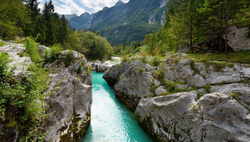 View of bright blue water running past giant stones with mountains in the background at Korita Soče (Great Soča Gorge).