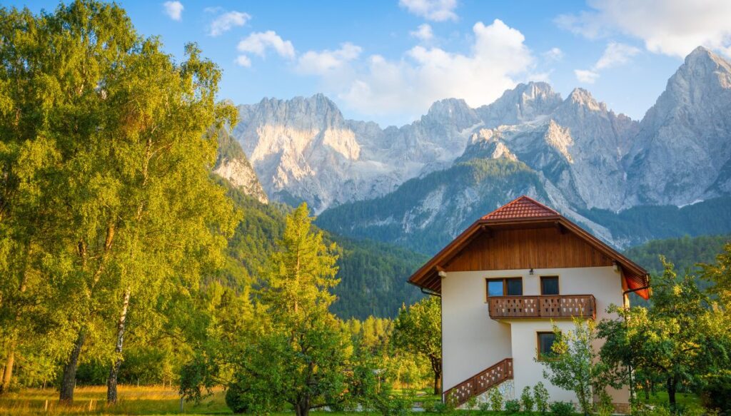 View of Kranjska Gora in the background. It is a mountains and you can see green trees on the left in the foreground and a building on the right that is alpine style.