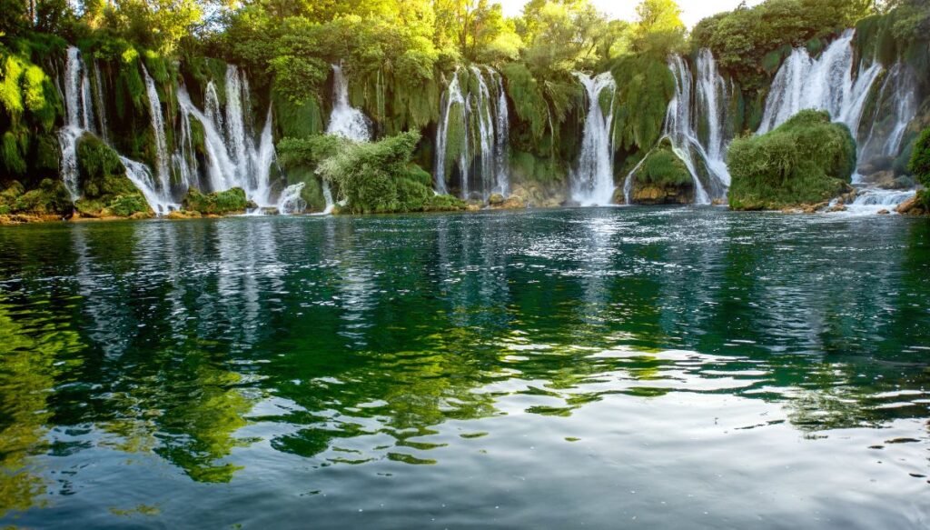 View of moss covered cliffs with water rushing down them into a beautiful blue pool. These are the Kravica Waterfalls.