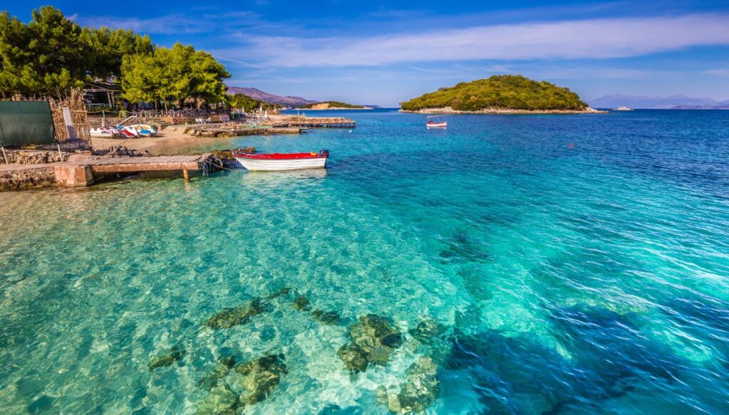 View of the clear blue water os the ocean near the shore of Ksamil. You can see a small white boat at the docks with trees and boats on the shore. There is also an island on the right.