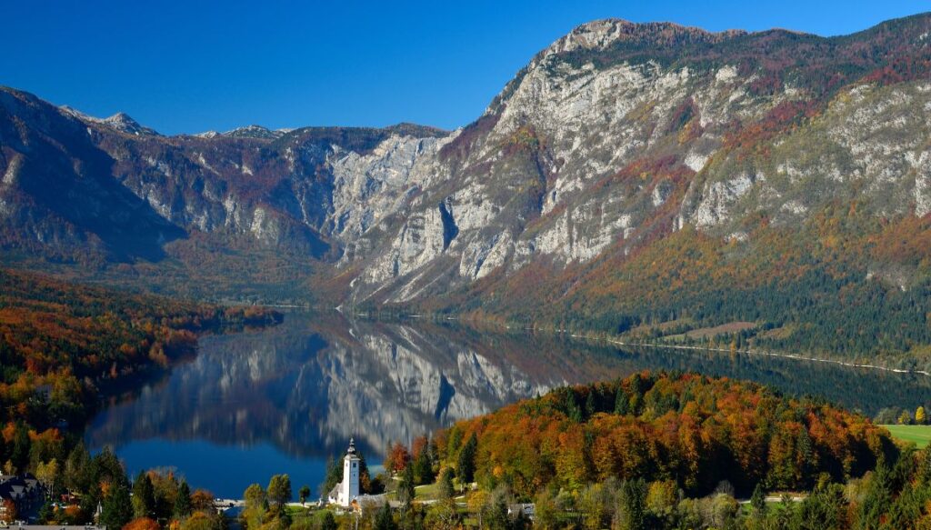 View of Lake Bohinj surrounded by mountains with trees changing color in the fall. You can also see a white church in the foreground on the edge of the lake.