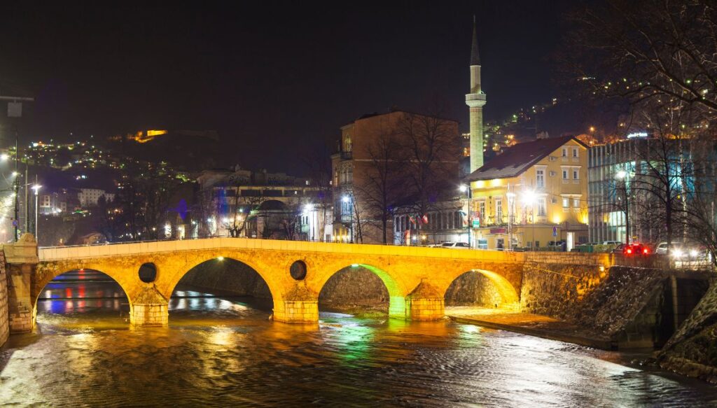 View of the arched Latin Bridge in Sarajevo lit up in the evening with houses and a mosque minaret all around it and a river running through the center of the city.