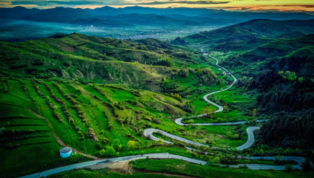 Aerial view of a road winding through green pastures with the sun setting in the background in Maramureș.