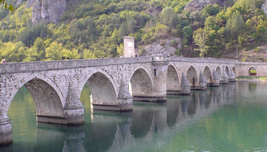 View of the arched, stone Mehmed Paša Sokolović Bridge spanning across bright blue water with people walking over it and trees all around.