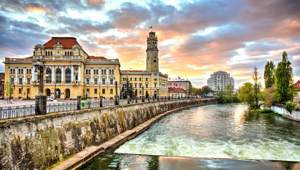 View of a giant Colonial yellow building on the left with a giant steeple and a river on the right with the sun setting in the background in Oradea.