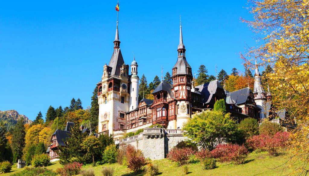 View of the beautiful spires of Peles Castle with a Romanian flag on top and trees surrounding the castle that are changing color in the Autumn.