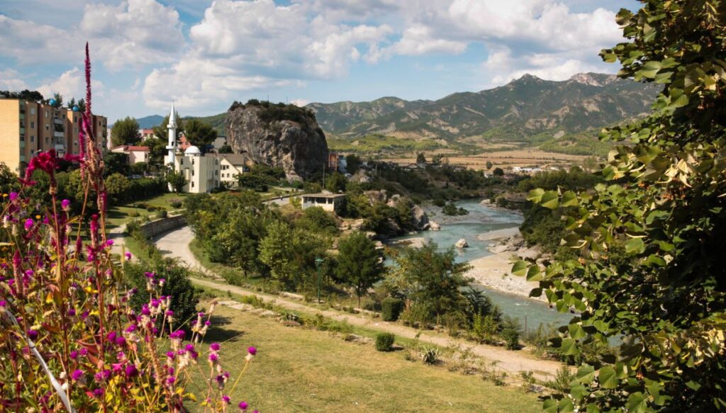 View of a path along the river with mountains in the background and a pink tree in the left foreground. You can see a town with a mosque on the left that is known as Permet.