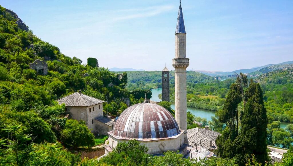 View of the minaret of a mosque surrounded by trees on a hill that overlooks a river with bright blue water in Počitelj.
