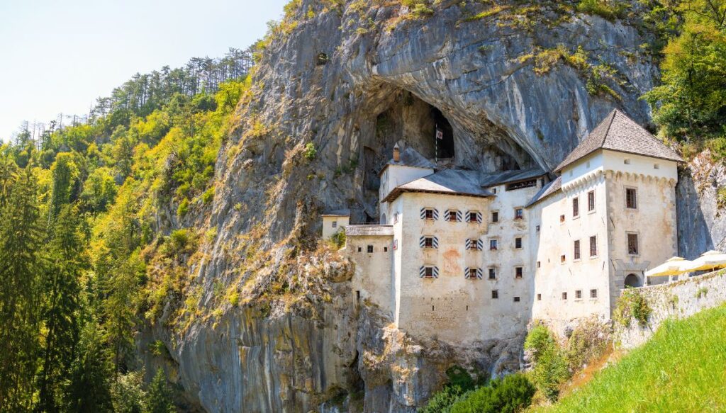 View of the white, stone Predjama Castle built into the rocks on the side of a mountain in Slovenia.