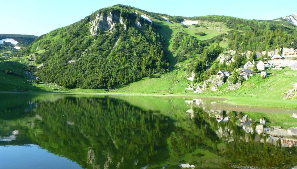 View of green mountains surrounding Prokoško Lake on a sunny day with wooden houses along the edge of the lake.