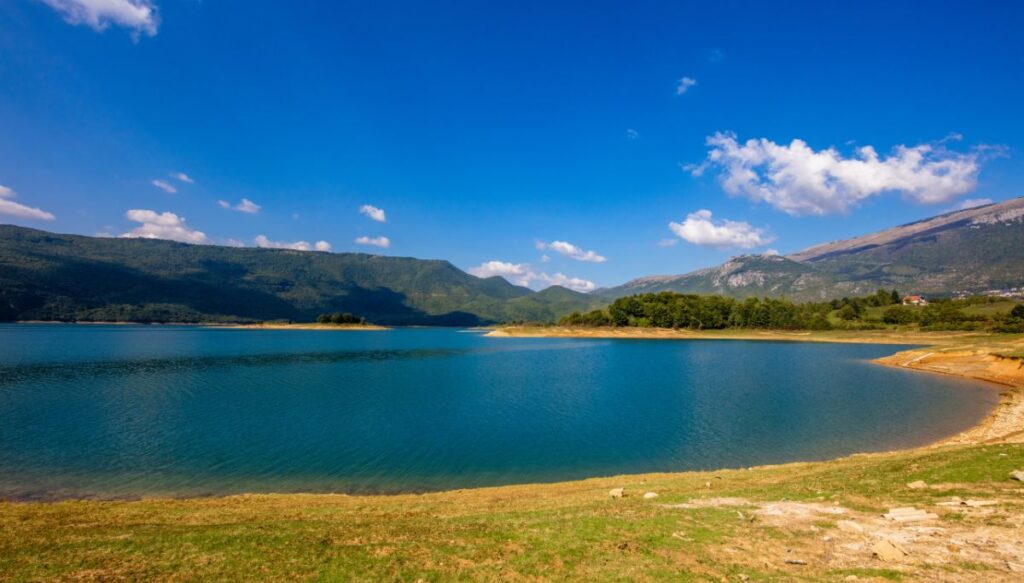 View of the bright blue water of Ramsko Lake sitting in the middle of the picture surrounded by green mountains and yellow grass.