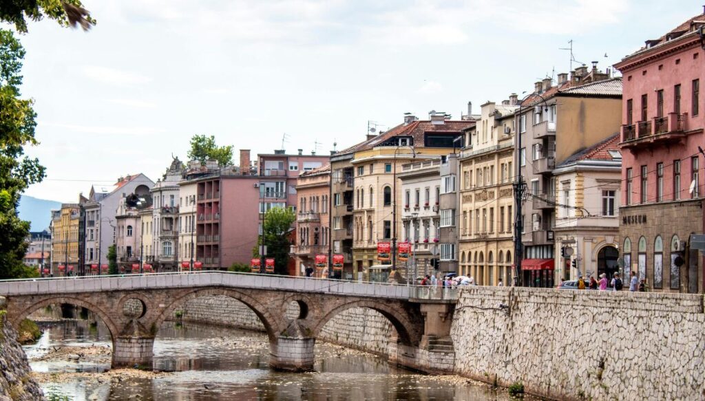 View of Colonial-style homes sitting along a stone wall on the river with a stone bridge going over the river in Sarajevo.