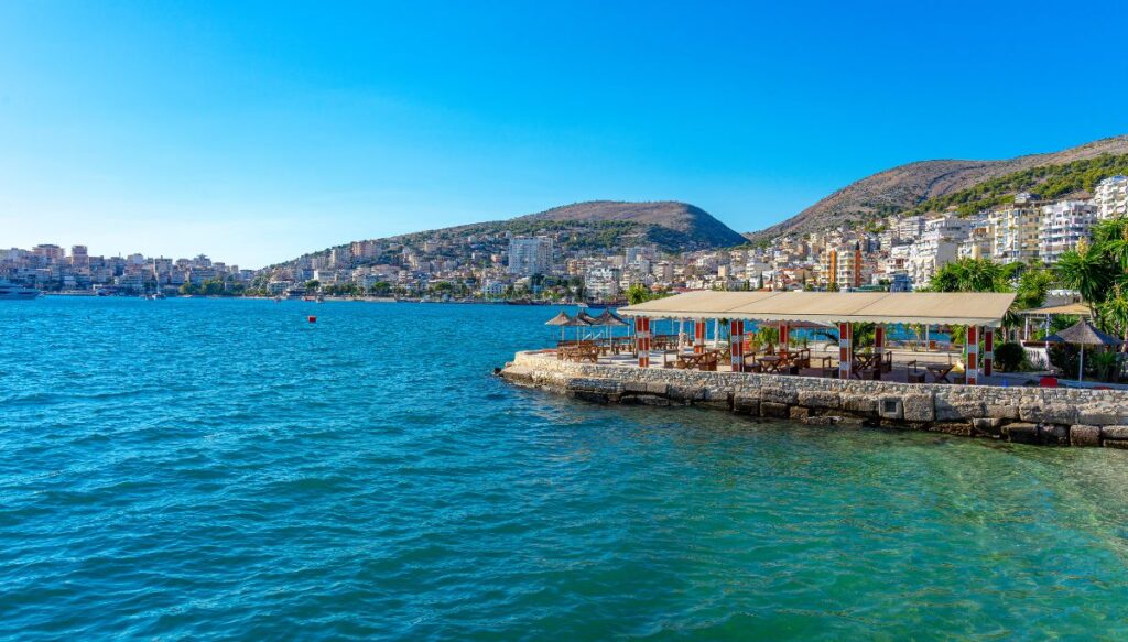 View of the blue waters in the Albanian Riviera and the builds along the coast with a restaurant along the water and surrounded by a stone wall on the front right.