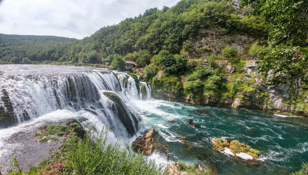 View of the Štrbački Buk waterfall that descends into a river full of bright blue water.