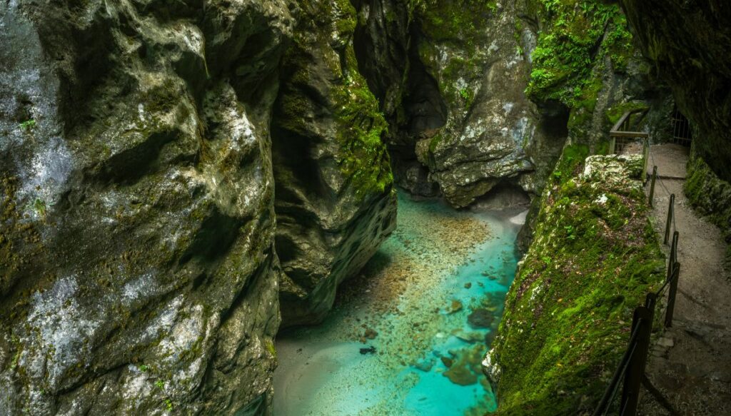 View of bright blue, shallow water running past large rocks covered in moss with a walking path with a fence on the right in Tolmin Gorge.