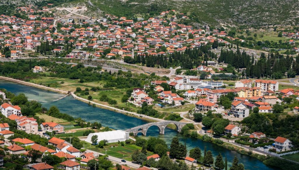 Aerial view of the red roofed homes in Trebinje. You can see the homes sitting in a valley with a blue river running through it and a stone bridge going over the river.