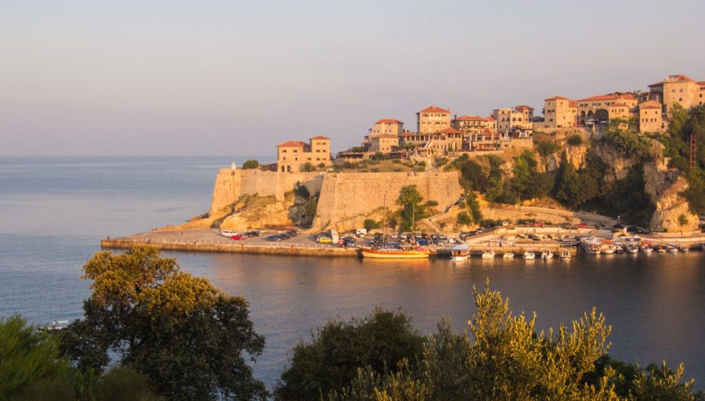 View of the town of Ulcinj at sunset from across the water. You can see houses on a hill with stone walls that lead down to a parking lot by the water. 