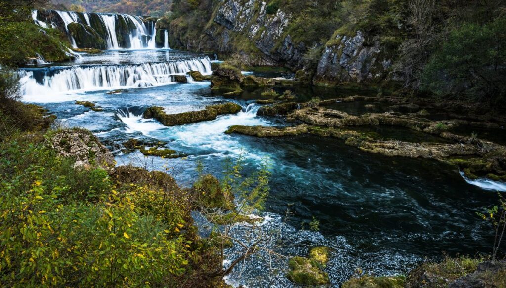 View of Waterfall Štrbački buk in Una National Park with the river running beneath the waterfall with bushes and trees all around it.