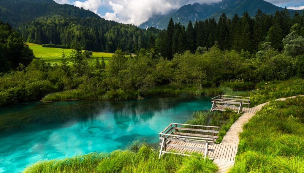 View of two wooden platforms sitting along the bright blue water of the river in Zelenci Nature Reserve with mountains all around it.