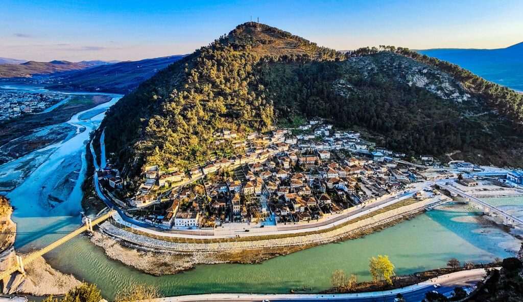 Aerial view of the mountains and white homes along the river in Berat with the sun setting in the background in one of the most beautiful places in Albania.