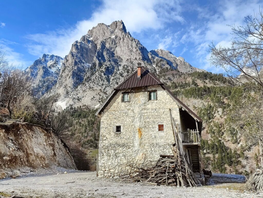 View of a stone house sitting in front of the Albanian Alps covered in snow in Valbonë Valley National Park.