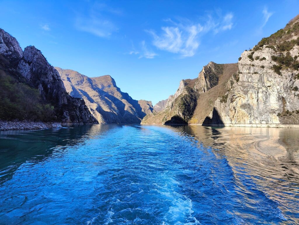 View of the bright blue waters of Lake Komani with giant mountains on either side.