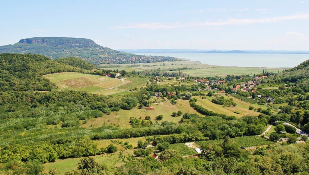 Aerial view of forests and meadows along the edge of Balaton Lake with houses in the distance and the lake in the distance at Balaton Uplands National Park.