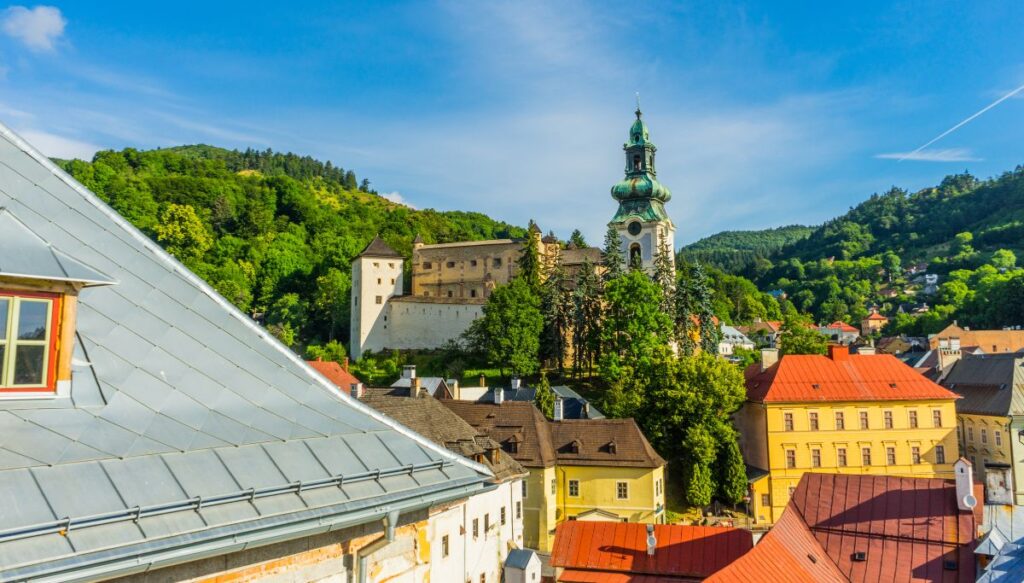 View of a church steeple from a rooftop that is surrounded by trees and vibrant buildings in Banská Štiavnica, Slovakia.