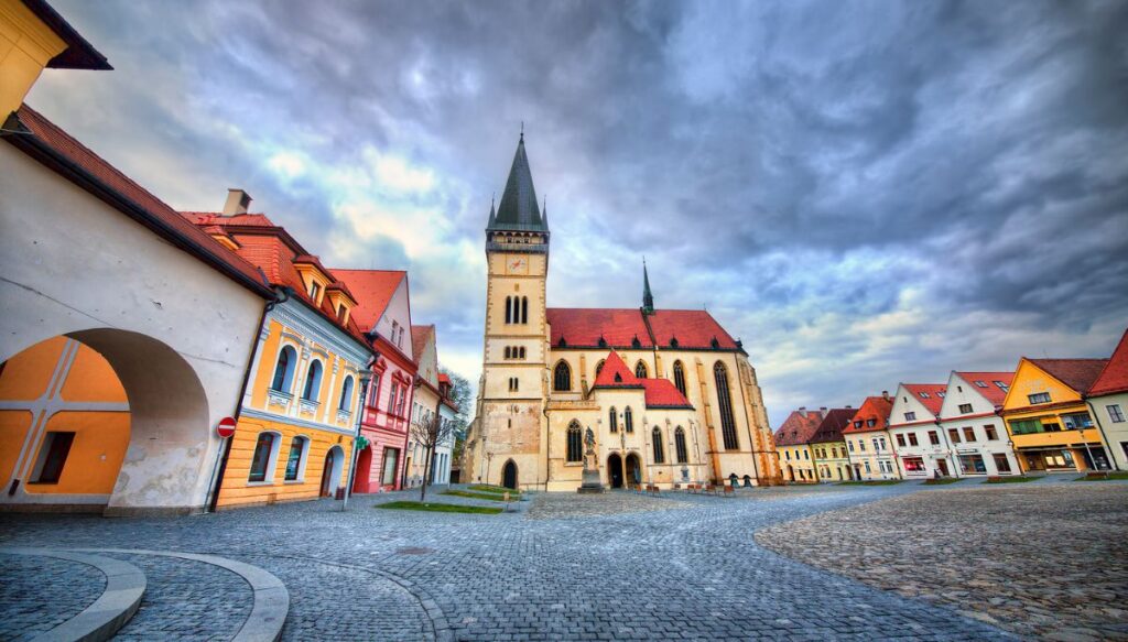 View of a large white church in the center of a square in Bardejov surrounded by vibrant buildings and sitting beneath a cloudy sky.