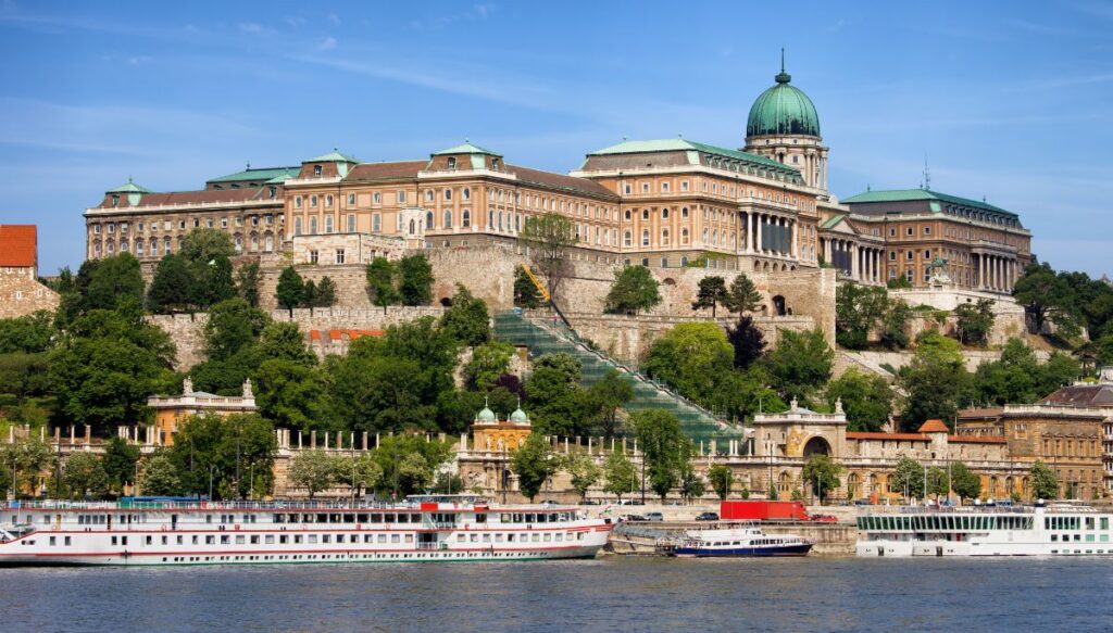 View of Buda Castle sitting on the top of the hill in Budapest with boats sitting along the edge of the Danube River in the city.